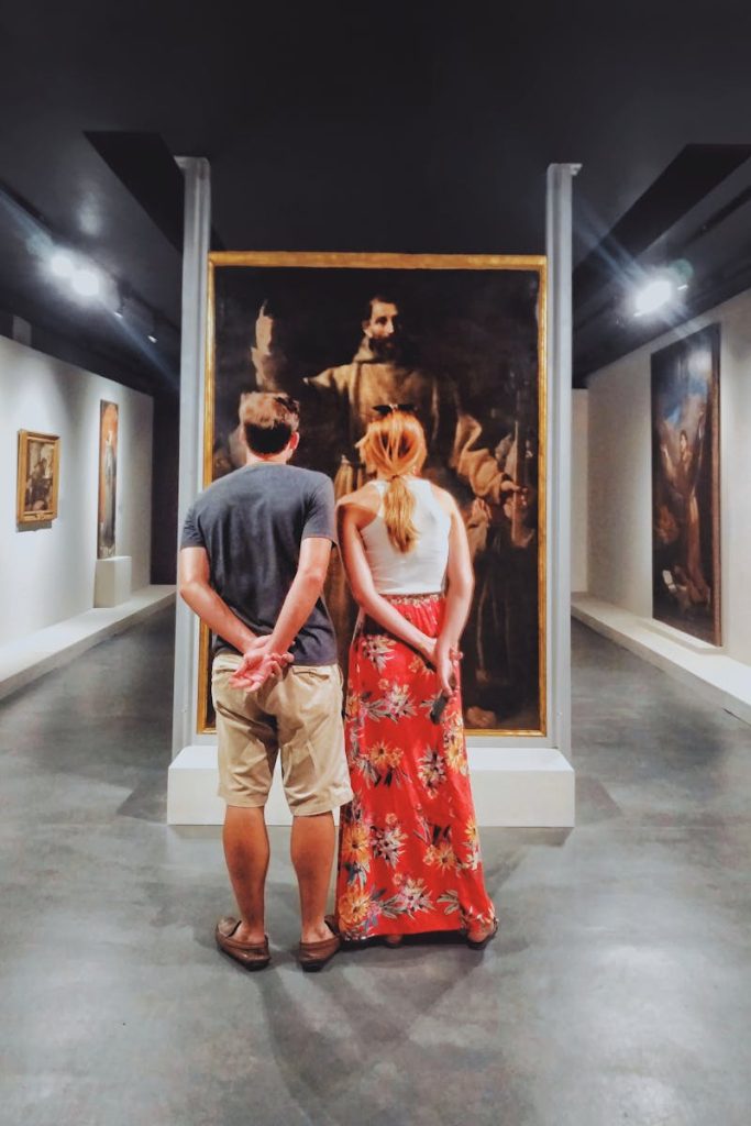 A couple stands inside an art gallery in Brazil, engaging with classical paintings.